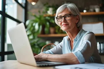 Fotobehang Smiling mature middle aged businesswoman using laptop working on sitting at desk. Happy old businesswoman hr holding cv interviewing distance applicant, senior seeker searching job, Generative AI © Hitesh