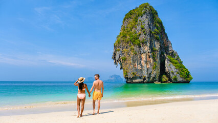 Fototapeta na wymiar a diverse couple of men and women on the beach, an Asian woman in bikini and a caucasian man in swim short walking on the beach of Railay Beach in Thailand with limestone cliffs in the background