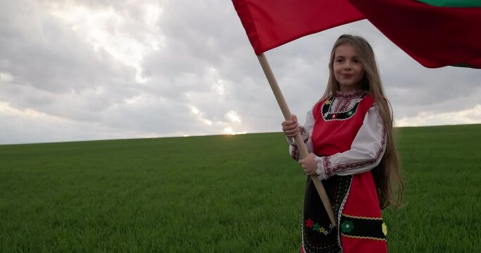 Bulgarian happy girl in ethnic folklore costume proudly waving bulgarian flag against sunset sky over field, 3th March Liberation day of Bulgaria