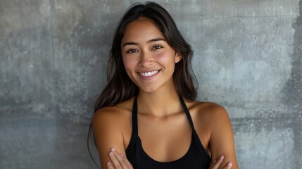 A portrait of a young Latin woman with a warm smile and arms crossed, standing against a grey wall
