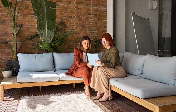 Two happy busy business women talking using tab working together sitting on sofa in green cozy office. Professional female colleagues employees looking at digital tablet discussing project at work.