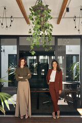 Two happy confident professional business women of young and middle age, female entrepreneurs or executive leaders working together standing at work in office looking at camera. Full length vertical.