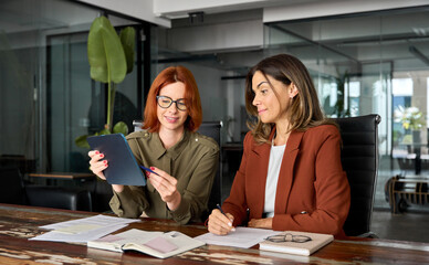 Two busy business women talking using tab working together at desk in office. Professional female...