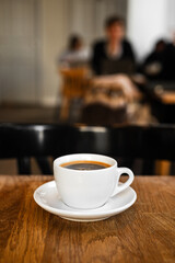 A white cup of black coffee on a saucer on the wooden table in a cafe, people in bokeh - 744464882