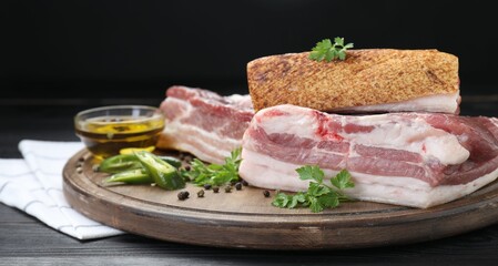 Pieces of raw pork belly, chili pepper, peppercorns, oil and parsley on black wooden table, closeup