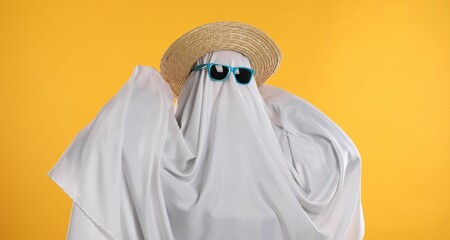 Person in ghost costume, sunglasses and straw hat on yellow background