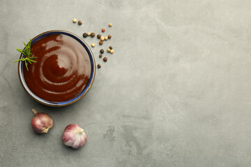 Tasty barbeque sauce in bowl, rosemary, garlic and peppercorns on grey textured table, flat lay. Space for text