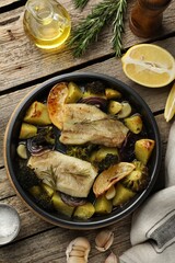 Pieces of delicious baked cod with vegetables, lemon and spices in dish on wooden table, flat lay