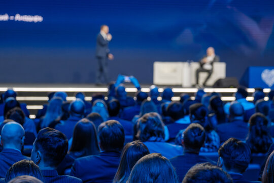 Out-of-focus shot capturing the atmosphere at a corporate event, with speaker presenting to an attentive audience.