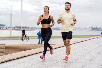 Fototapeta premium Couple in sportswear runs together on a city promenade with a marine backdrop.