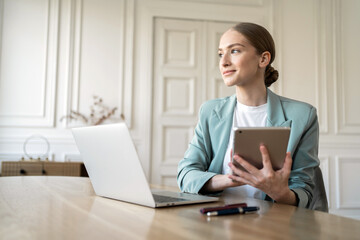 Professional woman contemplates while holding tablet, with laptop on desk.