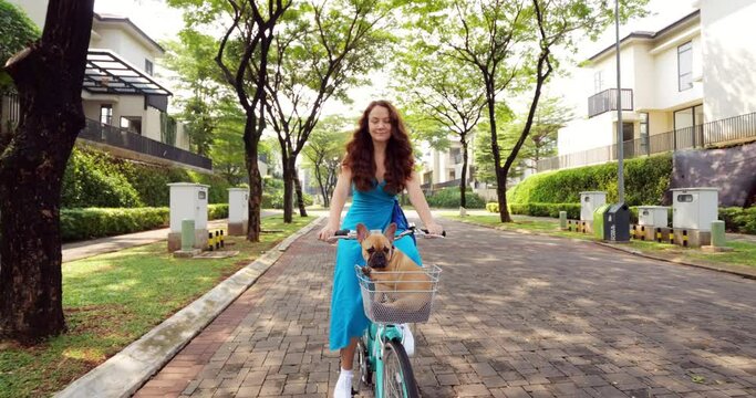 Happy young woman rides her bicycle on suburban street, accompanied by her beloved French Bulldog puppy in handlebar basket. Slow-motion reverse dolly shot. They cruise leisurely down empty alley