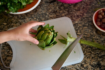 Close-up. Directly above human hands cutting fresh ripe artichoke on a cutting board. Healthy...
