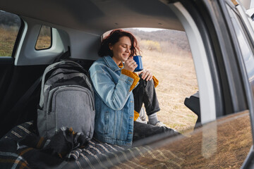 A young happy woman sitting in an open car trunk drinking tea from a mug. Travel by car concept - 744519267