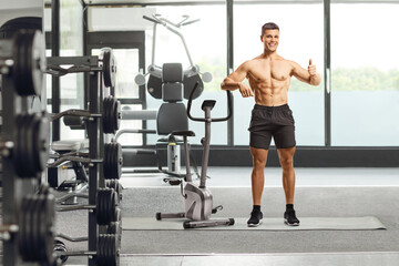 Fit man leaning on an exercise bike and gesturing a thumb up sign at a gym