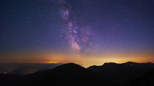 Time lapse of the Milky Way, the silhouette of the mountain landscape, the night city shining in the valley