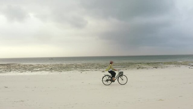 4K footage of young woman riding on bicycle along beautiful packed damp sand beach. Spending time outside, sports, leisure activities concept.. Environment, healthy lifestyle, outdoors concept.