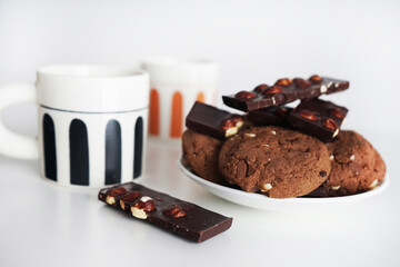 Two cups of tea next to chocolate chip cookies and chocolate with nuts on a plate on a white background