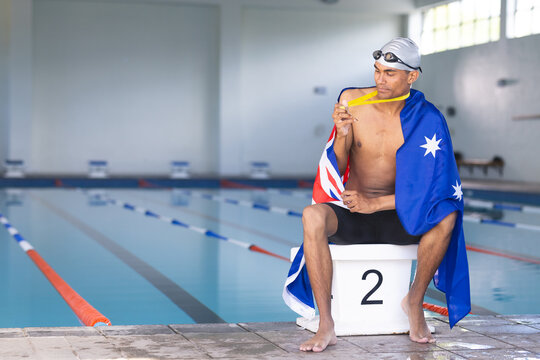 Young biracial male swimmer wrapped in an Australian flag sits poolside with a medal