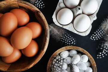 Brown eggs in a wooden bowl, white chicken eggs in a tray next to quail eggs and feathers on a dark...