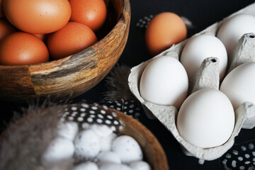 Brown eggs in a wooden bowl, white chicken eggs in a tray next to quail eggs and feathers on a dark background