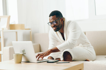 African American man working on a laptop in his modern home office, wearing a bathrobe and sitting on a sofa He is typing with a smile on his face, enjoying a productive morning in the cyberspace of