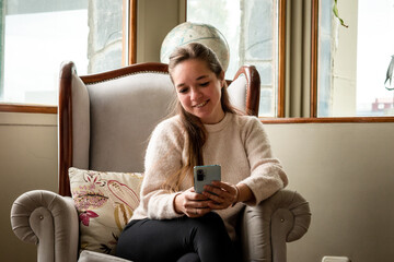 Mujer viendo su teléfono móvil sentada en el sofá de su sala de estar	 - obrazy, fototapety, plakaty