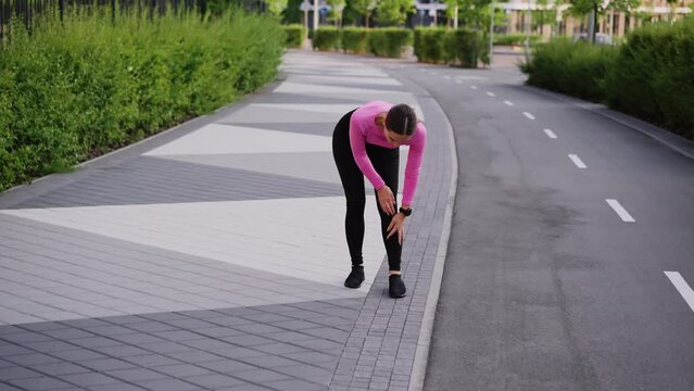 A young sportive woman stops jogging, got injured of leg and feet, has cramp, sprain or dislocation. An athlete girl stands on the sidewalk, wrinkles face in pain, steps, limps and holds knee. The