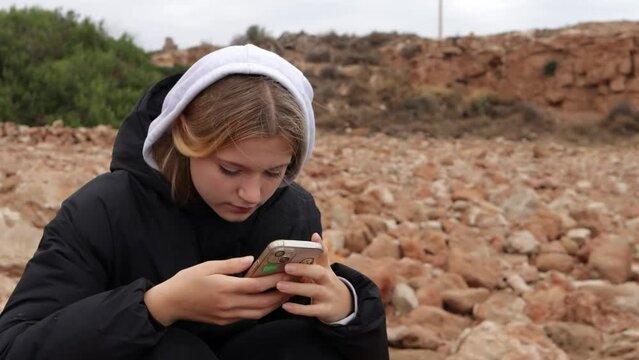 An attractive young girl stands on a rock on the seashore and looks at her phone. A Caucasian blonde stands on the seashore with a phone in her hands. A teenage girl in a jacket sits on the seashore.