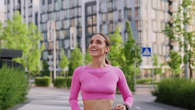 A young sportive girl jogger outdoors in the city park among modern residential highrise buildings. A happy smiling sportswoman shows perfect running technique. The concept of only forward to victory