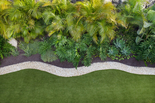 Aerial view of a meticulously landscaped tropical garden, featuring lush tropical trees on one side and a pristine stretch of green lawn on the other, divided by a border of decorative white stones