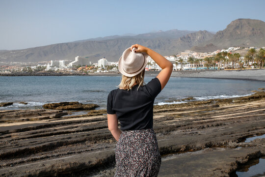 Rear view of young Caucasian woman wearing dark colors and holding her hat with one hand while looking towards the coast of Las Americas, favorite tourist destination, Tenerife, Canary Islands, Spain