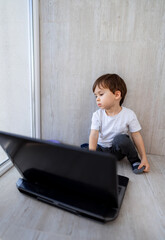 Little boy sits on the floor and looks at the laptop. The child is sitting in front of the computer and playing on the Internet.