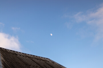 Fototapeta na wymiar Moon in the blue sky with white fluffy clouds and roof on the foreground