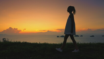 Woman enjoy aerial sunset on tropical island hill. Girl silhouette walking green grass meadow, colorful orange sky in background. Beauty nature. Outdoor lifestyle travel on summer holiday vacation