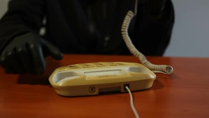 An unknown man wearing black gloves dials a number and calls on a landline phone.