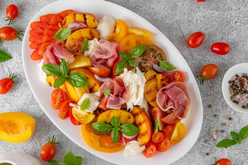Caprese salad with mozzarella, tomatoes, prosciutto and grilled peaches on a white plate on a gray concrete background. Fresh summer salad. Top view.