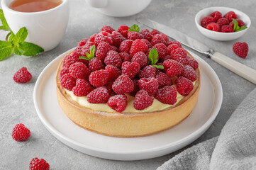 Delicious raspberry tart with vanilla custard cream sprinkled with powdered sugar on a white plate on a gray concrete background. Summer dessert.