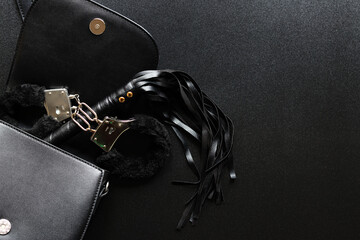 Sex toys. A small black bag with game handcuffs and a leather whip for intimate games. Women's objects for sexual entertainment on a black background - obrazy, fototapety, plakaty