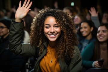 A cheerful young woman with curly hair raising her hand to participate during a lecture in a classroom setting, showcasing active engagement and a positive attitude