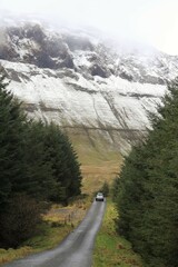 Car travelling along country road bordered by forest, towards snow-covered mountain cliff face shrouded in cloud. Gleniff Horseshoe valley, County Sligo, Ireland