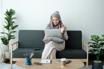 Beautiful asian woman in hijab works inside a modern office building, Muslim talks on video call, online meeting with colleagues and clients, woman cheerfully waves her hand and greets interlocutors