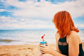 Beautiful woman in swimsuit is sitting on beach with cocktail and look at ocean. Tropical alcohol beverage. Summer day with cloudy sky. Red-haired lady with sunglasses on vacation. Holidays at sea.