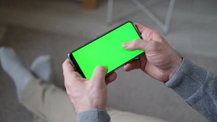 Young man sitting at home holding a green smartphone screen mockup in his hand. A man plays online games on a chromatic mobile phone. Horizontal mode. Clicking, playing, surfing social networks