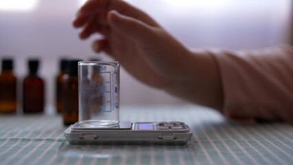 Closeup side view of female artisan puts transparent measuring cup on electronic scale to prepare mixture for handmade scented candle. Process of making handmade natural scented candle at workshop.