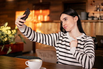 Photo of young woman drinking coffee while taking selfie on smartphone in cafe indoors