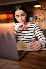 Woman in earphones using laptop while sitting at cafe. Young female student sitting in a coffee shop and working on laptop, looking conference.