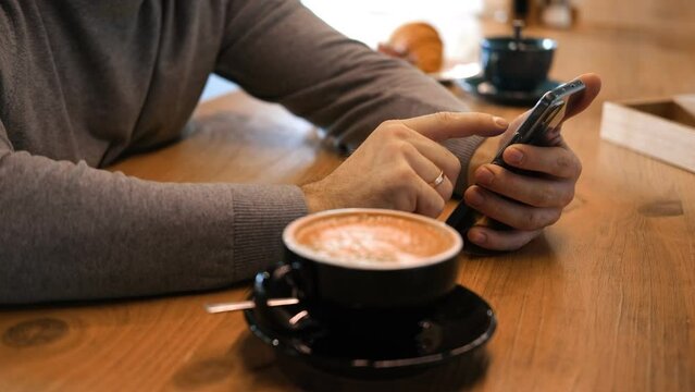 Adult Man Wearing Grey Sweater Set at Wooden Table in a Cafe Using Smartphone. Freelancer's Cafe Break. Stylish Male Enjoying Coffee and Phone Time in Cozy Coffee Shop Atmosphere. Freelancing, Leisure