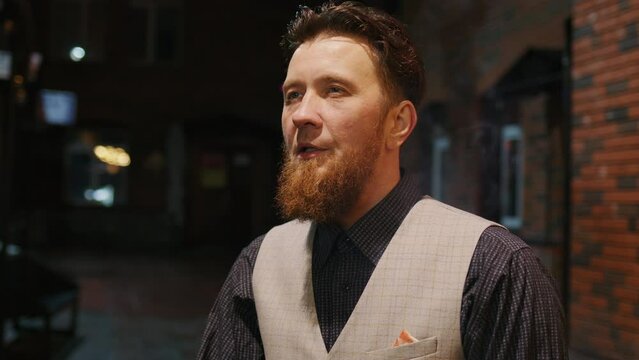 Red-haired middle-aged man having cigarette in yard of brick house. Bearded guy takes puff and lets out smoke, standing on dark street. Smoker doing smokebreak, thinking about something outdoor