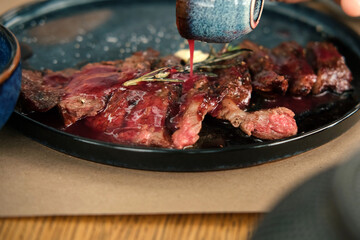 Man Cutting Rib Eye Steak, Pouring it Red Sauce on a Black Plate. Person Having a Delicious Lunch. Grilled Sirloin Beef Steak with Rosemary and Garlic on a Wooden Table. BBQ Meat Delicacy Bio Food.
